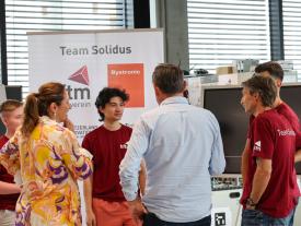 Personen diskutieren vor einem Poster von Team Solidus.